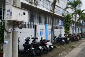 Tòa nhà nhiều tầng với dãy xe máy đỗ phía trước và hàng cây dọc lối đi