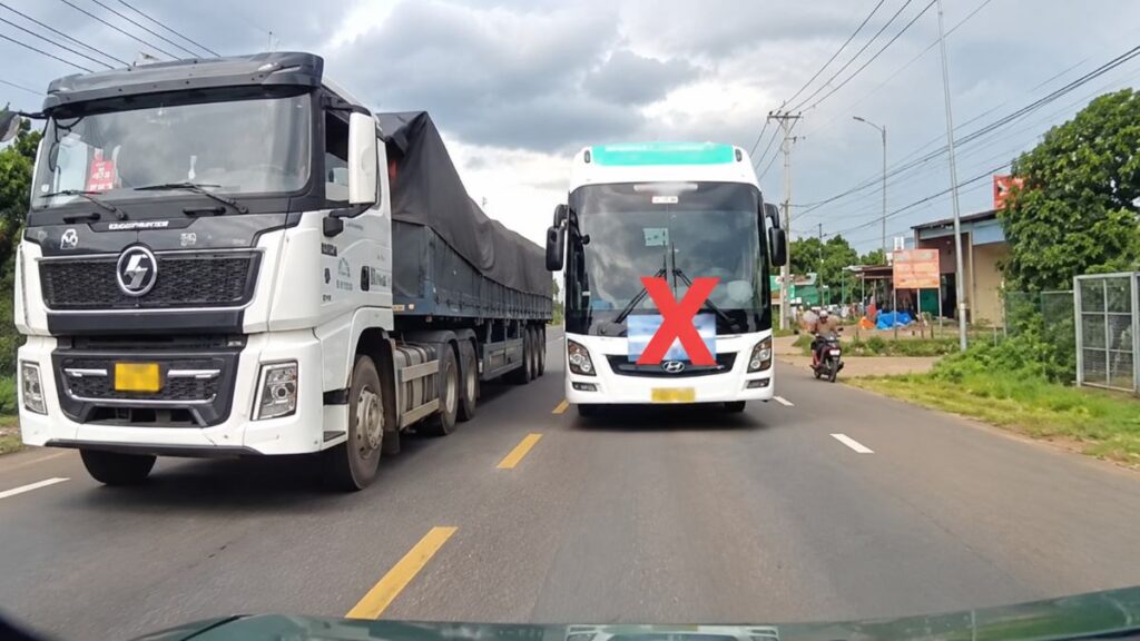 Xe khách màu trắng đi đối diện xe tải lớn trên đường hai chiều, phía trước xe khách có dấu X màu đỏ.