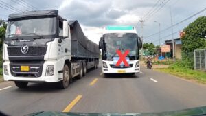 Xe khách màu trắng đi đối diện xe tải lớn trên đường hai chiều, phía trước xe khách có dấu X màu đỏ.